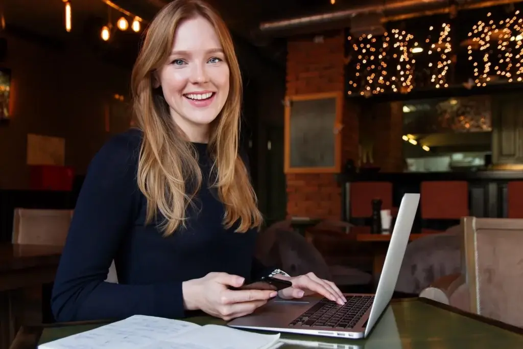 "Eine lächelnde Frau mit langen blonden Haaren sitzt in einem Café an einem Tisch, hält ein Smartphone in der Hand und arbeitet an einem Laptop, umgeben von Notizen, symbolisch für Jobcoaching in Magdeburg und berufliches Wiedereinsteigen nach einer Pause.